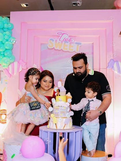 A happy family celebrating a second birthday with a cake cutting ceremony in front of a donut themed backdrop. The scene is filled with colorful props and a clown entertainer.