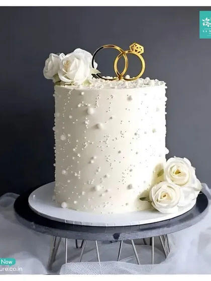Simplicity at its finest. This tall, single-tier engagement cake is decorated with delicate sugar pearls and white roses, topped with an elegant intertwined rings topper, making it a perfect choice for a chic and modern celebration.