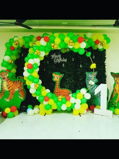 A jungle-themed first birthday party with a circular balloon arch against a green hedge backdrop. The animal cutouts and number '1' prop complete the theme.