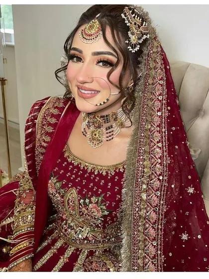 A stunning Pakistani-inspired bridal look. This style features dramatic winged eyeliner, a contoured base, and soft curls, all complementing her intricate red sharara and traditional jewelry.