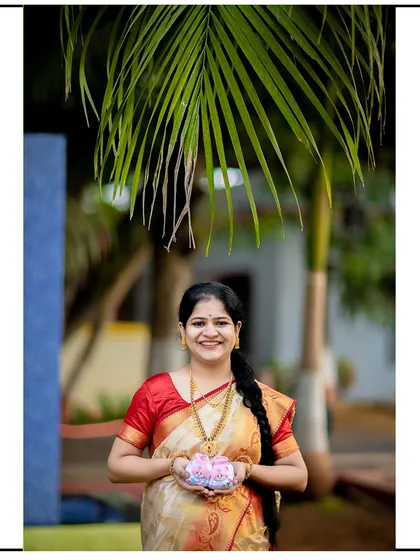 A happy mom-to-be in a traditional saree, proudly holding up a pair of pink baby booties.