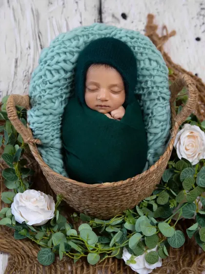 A classic newborn pose with a nature-inspired touch. The baby is swaddled in a deep green wrap and bonnet, nestled in a basket adorned with eucalyptus leaves and white roses.