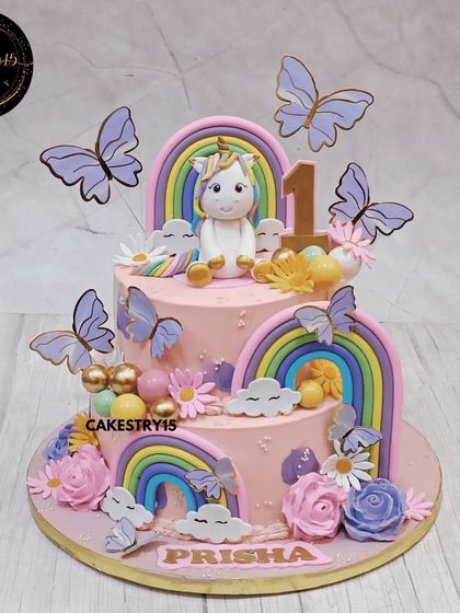 A beautiful two-tier pineapple cake in soft pink for a first birthday. The design is a fantasy of unicorns and rainbows, decorated with fluttering butterflies and colorful flowers.