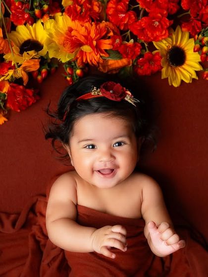 A sweet smile from a baby girl lying on a rich, rust-colored blanket. The warm tones and autumnal flowers create a cozy and elegant portrait.