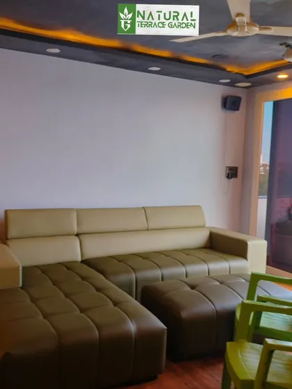 The interior of a compact and cozy terrace room, designed as a personal relaxation space. It features a comfortable L-shaped sofa and a foot massager, with a large window overlooking the terrace.