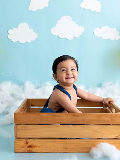 A happy baby sits in a wooden crate surrounded by clouds and balloons.