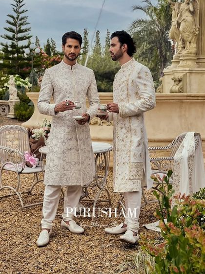 In the heart of elegance, two gentlemen savor a quiet moment. I designed these coordinated ivory sherwanis, one with geometric embroidery and the other with floral, to showcase individual style within a shared aesthetic.
