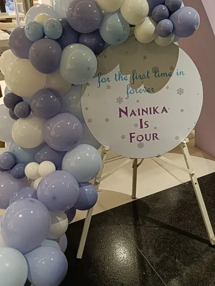 "For the first time in forever, Nainika is four!" This custom welcome sign for a Frozen-themed party is framed by a beautiful balloon arrangement in icy shades of blue, purple, and white, setting a magical tone from the start.