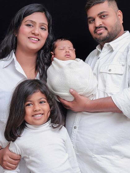 A bright and happy family portrait celebrating their new addition. Coordinated in white, the focus remains entirely on the family's smiles and the peacefully sleeping newborn held securely in dad's arms.