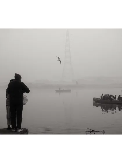 A black and white scene at the Yamuna Ghat, where two people feed the birds as a boat full of tourists passes by. The foggy atmosphere creates a layered and moody composition.