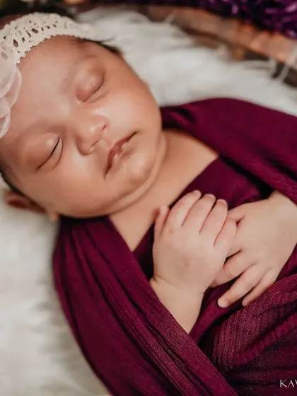 This sweet newborn is sleeping peacefully, adorned with a delicate floral headband. The deep magenta wrap and surrounding flowers create a rich, beautiful color palette.