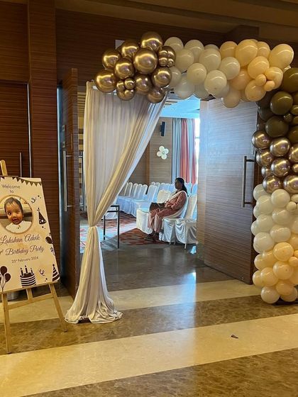 A simple and elegant entrance decoration for a first birthday party. The setup includes a welcome stand with the baby's photo and a beautiful organic balloon arch in muted gold, silver, and peach tones.