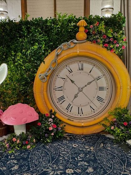 A fantastic photo corner featuring a giant yellow pocket watch prop against a green hedge wall. A white rabbit figure and illuminated butterfly add to the magical, wonderland feel.