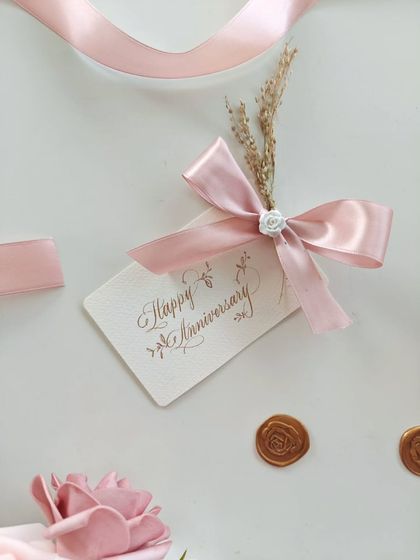 A close-up of the elegant "Happy Anniversary" gift tag, featuring delicate calligraphy and adorned with a pink satin ribbon and dried flowers.