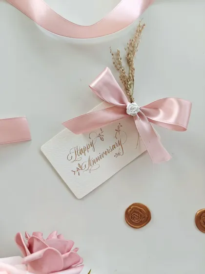 A close-up of the elegant "Happy Anniversary" gift tag, featuring delicate calligraphy and adorned with a pink satin ribbon and dried flowers.