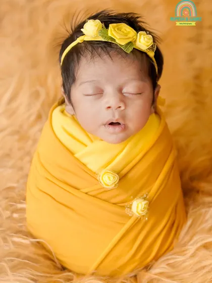 A sweet newborn girl in a yellow swaddle and matching floral headband, captured in a classic "potato sack" pose that is both safe and adorable.