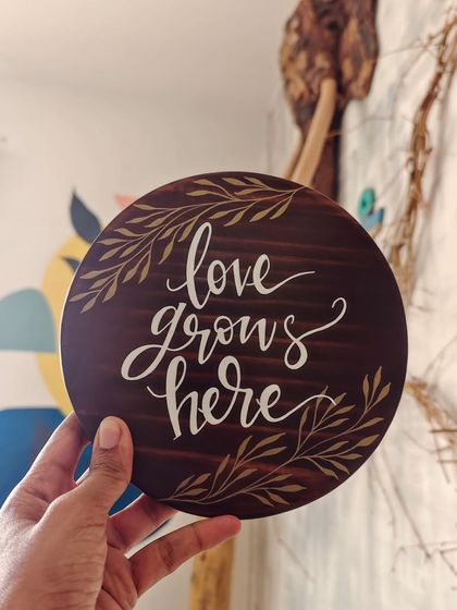 "Love grows here." This simple, elegant message is hand-painted on a round, dark-stained wooden plaque, perfect for celebrating a new home, a wedding, or the love in your family.