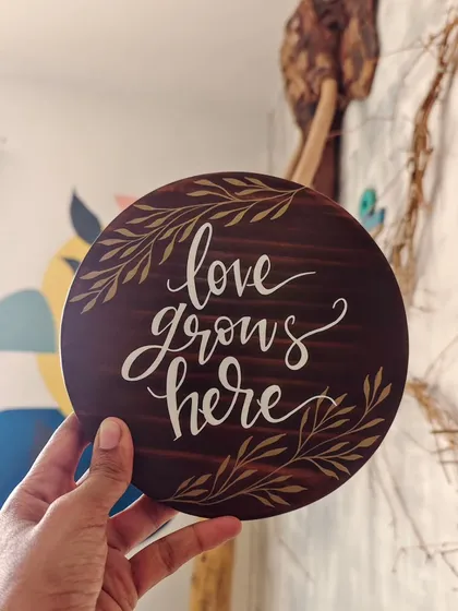 "Love grows here." This simple, elegant message is hand-painted on a round, dark-stained wooden plaque, perfect for celebrating a new home, a wedding, or the love in your family.