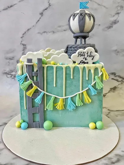 Celebrating a half birthday! This "halfway to one" cake is a creative square design with a hot air balloon topper, fluffy buttercream clouds, and a cute tassel garland made of fondant.