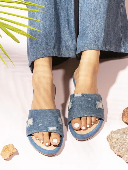 An on-model shot of distressed denim slides, styled with denim jeans. This shows the footwear in a casual, trendy, and relatable context.