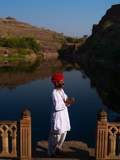 A man in traditional white clothes and a red turban stands in a moment of prayer by a lake. The tranquil setting provides a peaceful contrast to the usual bustle of Rajasthan.
