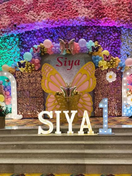 The main stage decoration for a butterfly-themed first birthday, featuring a sequin backdrop, large butterfly wings, marquee letters, and colorful lighting.