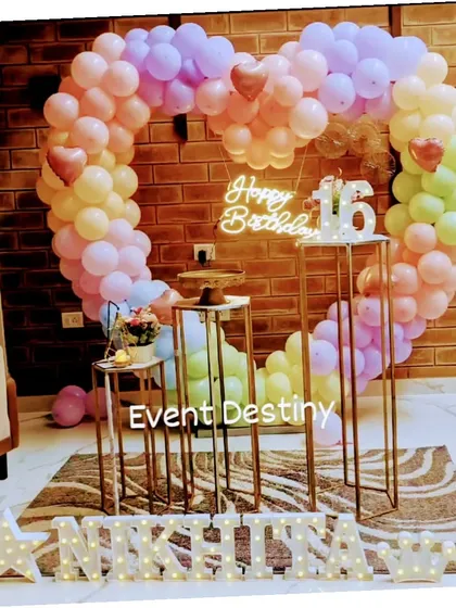 A sweet sixteen birthday setup in a client's home, featuring a large heart-shaped balloon arch in pastel rainbow colors, a custom neon sign, and marquee letters for the name.