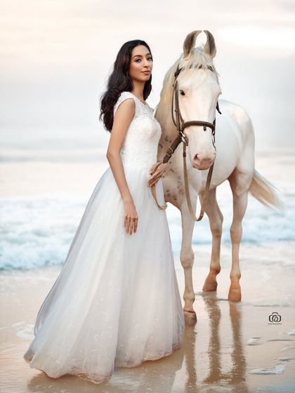 A wide shot of the model in a white gown leading the horse along the shoreline. This image tells a story and captures the magical, fairytale quality of the scene.