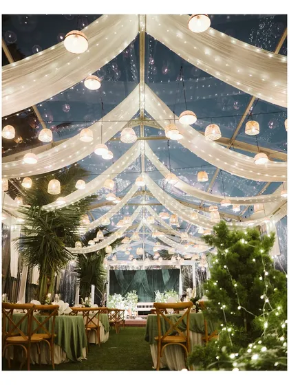 An enchanting evening reception setup under a clear tent adorned with fairy lights and flowing drapery. The lush greenery and warm lighting create a magical and inviting atmosphere for celebration.