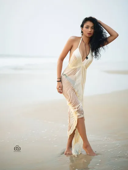 A three-quarter shot of a model in a flowing white dress, standing on the shoreline. The wind catches the dress, adding a sense of movement and life to the portrait.