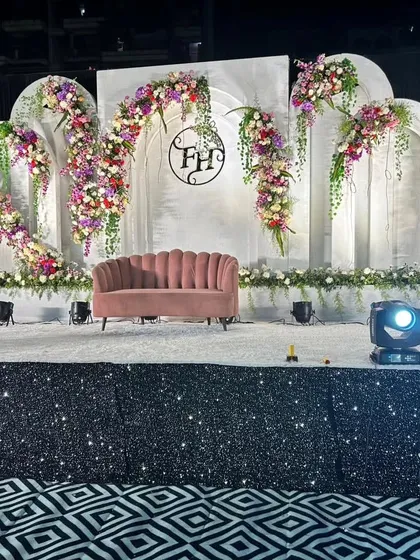A grand and elegant reception stage we designed, featuring a pristine white backdrop with classic architectural arches. The look is softened with cascading floral arrangements in purple, red, and white, and a custom monogram for the couple, creating a personalized and sophisticated focal point.