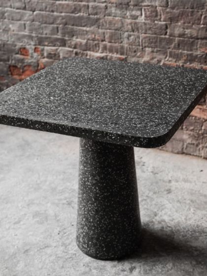 An angled view of the dark terrazzo cafe table, showing its sleek silhouette against a rustic brick wall. A perfect blend of industrial and modern design.