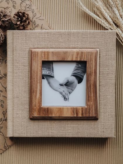 The cover of a rustic, linen-bound wedding album with a wooden frame inset, holding a photo of the couple holding hands.