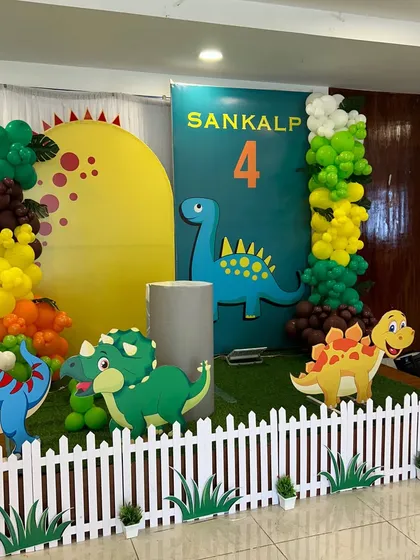 A friendly Dinosaur theme party with cute cartoon dinosaur cutouts, a white picket fence, and a colorful balloon arrangement.