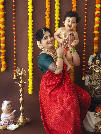 A mother in a vibrant red saree lifts her laughing baby, creating a dynamic and joyous portrait against a traditional backdrop.