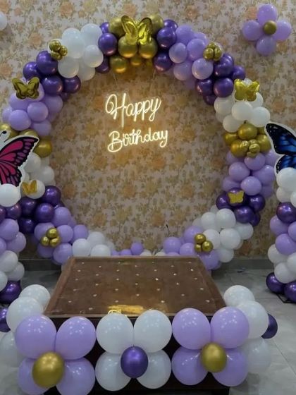A butterfly-themed birthday party with a purple and white balloon ring. The setup is accented with gold butterfly cutouts and a 'Happy Birthday' neon sign.