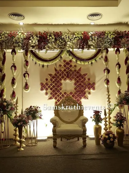 An elegant single-seater setup for a traditional ceremony. The ornate chair is placed before a backdrop decorated with a geometric floral pattern and hanging garlands.