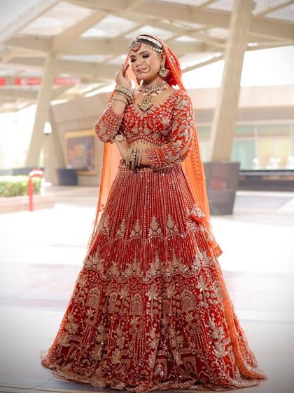 A full-length shot of a bride in a stunning orange-red lehenga. The makeup features a warm-toned palette to perfectly match the outfit.
