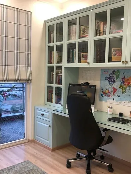 We designed this study area within a bedroom, featuring a custom-built unit with glass-fronted cabinets for books and a spacious desk. The setup is positioned by a large window with roman blinds, allowing for plenty of natural light. The balcony access provides a refreshing break space.
