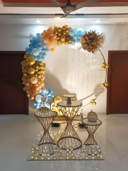 A semi-circular balloon frame in blue, gold, and peach, arranged with decorative tables for a modern birthday setup at home.