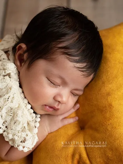 A close-up of a one-month-old baby sleeping on a tiny yellow pillow. The lace blanket adds a touch of delicate texture to this warm and sunny portrait.