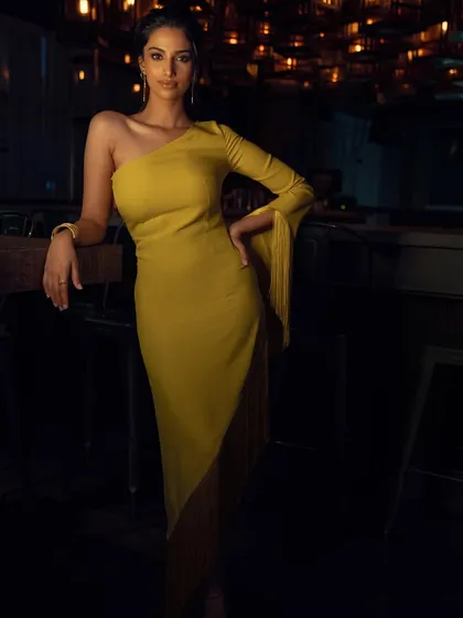 A full-length shot of a stunning yellow one-shoulder dress with fringe details. The styling is modern and sophisticated, set against a moody, ambient background.