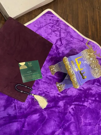 This personalized gift set features a vibrant purple prayer mat paired with a custom resin Quran holder. The holder is decorated with gold glitter and pearls, and the set includes a matching tasbeeh.