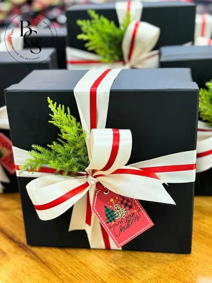 A classic Christmas gift box, elegantly wrapped in black and tied with a red and white striped ribbon. A sprig of fresh cypress and a festive "Merry Christmas" tag add the perfect touch of holiday spirit.