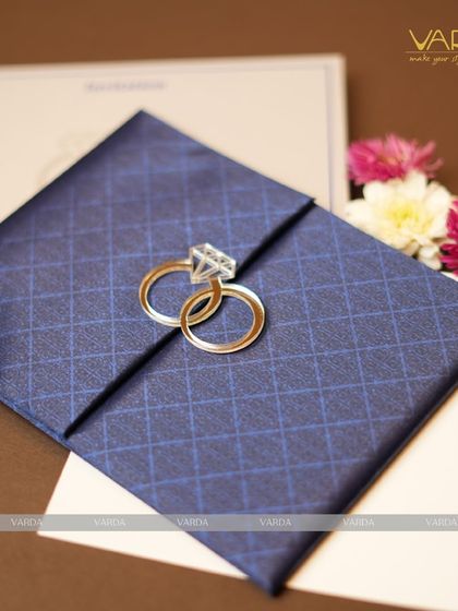 This engagement invitation features a deep blue, diamond-patterned textured folder held together by a unique clasp of two interlocking rings. The design is modern, stylish, and symbolic of the couple's union.