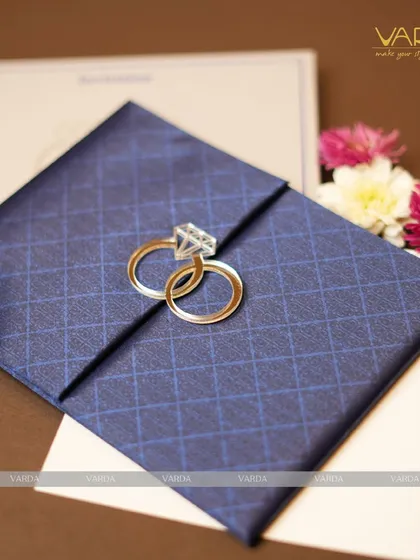 This engagement invitation features a deep blue, diamond-patterned textured folder held together by a unique clasp of two interlocking rings. The design is modern, stylish, and symbolic of the couple's union.