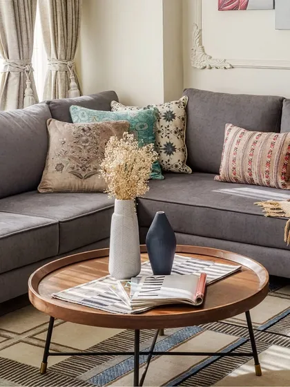 A cozy corner perfect for unwinding. The grey sectional sofa is layered with pillows in varied patterns and a soft throw, while the round wooden coffee table holds dried flowers, adding a natural, rustic touch.