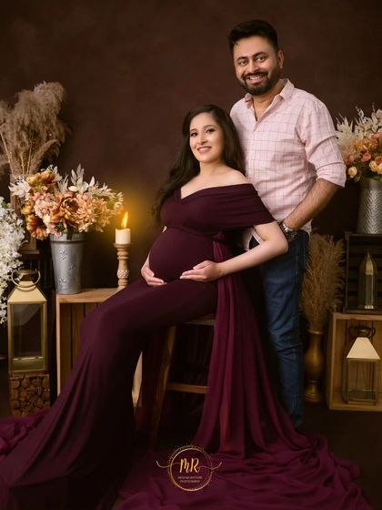 A classic, smiling portrait of an expecting couple. They pose together in a rustic studio setting, looking happy and excited for their new arrival.