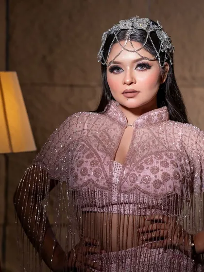 A model posing in a beautiful onion-pink gown with fringe details on the sleeves and a unique headpiece.