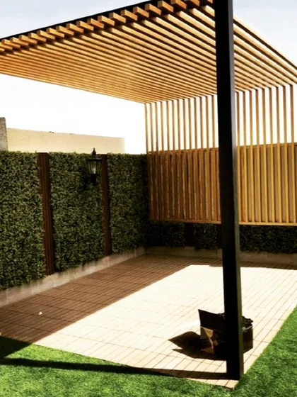 A modern pergola design combining a metal frame with a wooden slatted roof and side screen for partial privacy. The structure is set on a tiled patio next to an artificial turf lawn and a green wall.